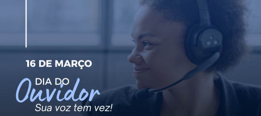 Uma mulher sentada, usando um headset de atendimento, em frente a um computador, olhando para o lado e sorrindo.