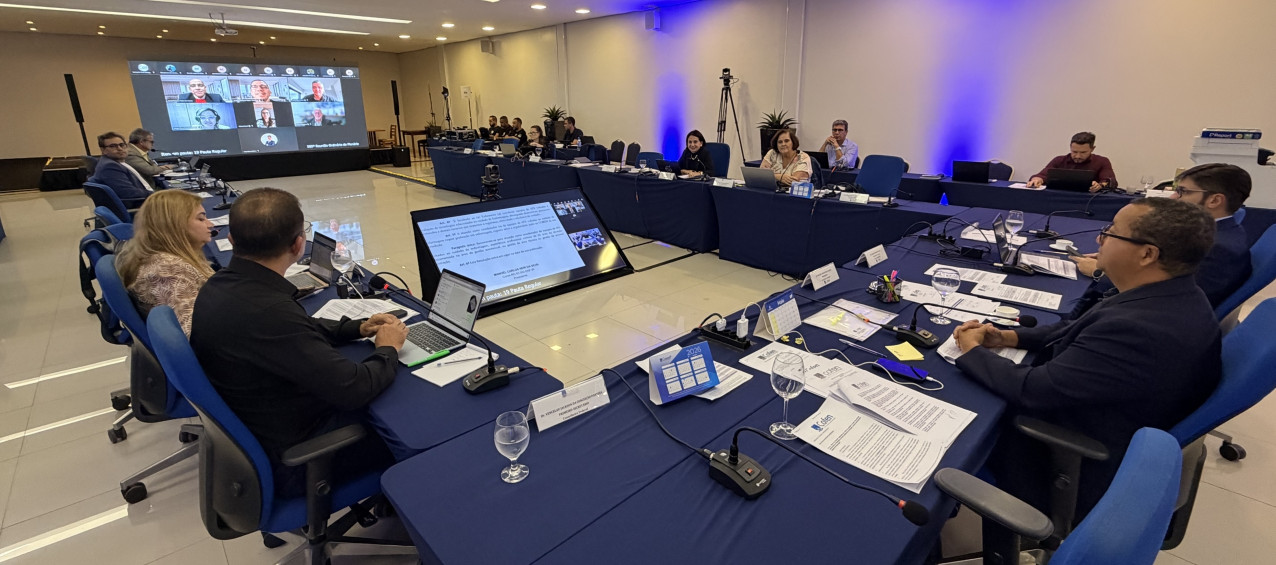 Reunião em sala com homens e mulheres sentados ao redor de uma mesa, enquanto um telão exibe participantes conectados virtualmente.