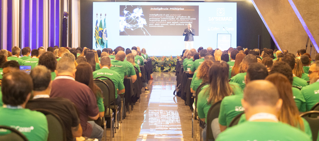 Pessoas sentadas em um auditório e uma pessoa falando no palco
