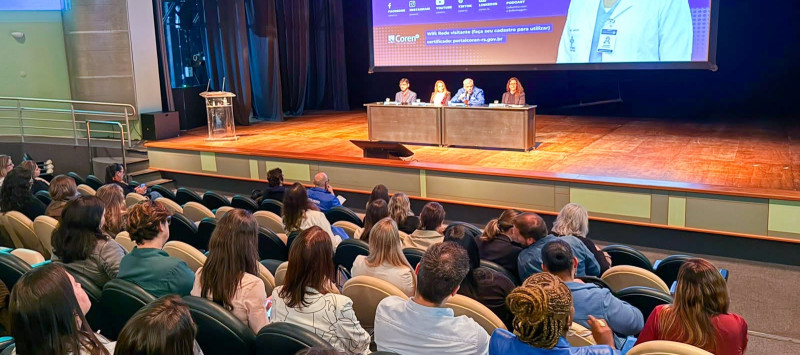 Auditório com mesa composta por autoridades à frente e público acompanhando o evento.