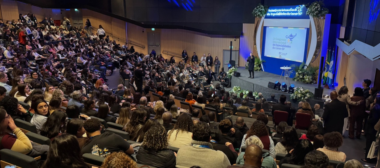Um homem falando ao microfone em um evento. Atrás dele tem outras pessoasl